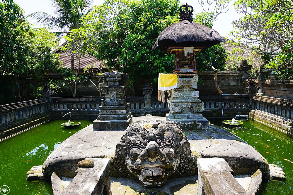 Intricate stone sculpture at Puseh Batuan temple, Bali, surrounded by lush greenery and water.