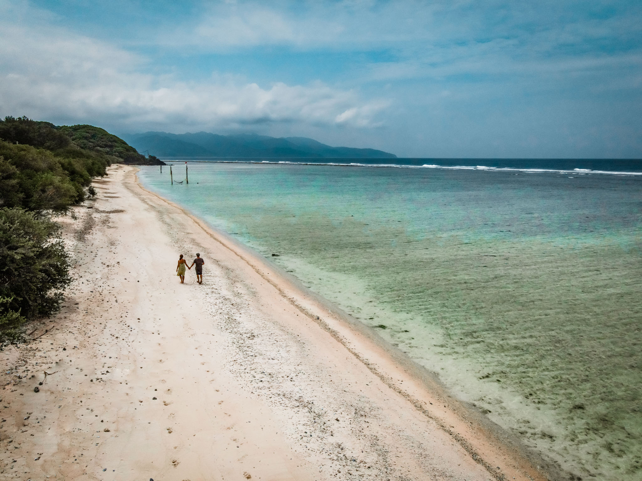 Couple walking on a secluded beach in the Gili Islands, with blue water and lush greenery.