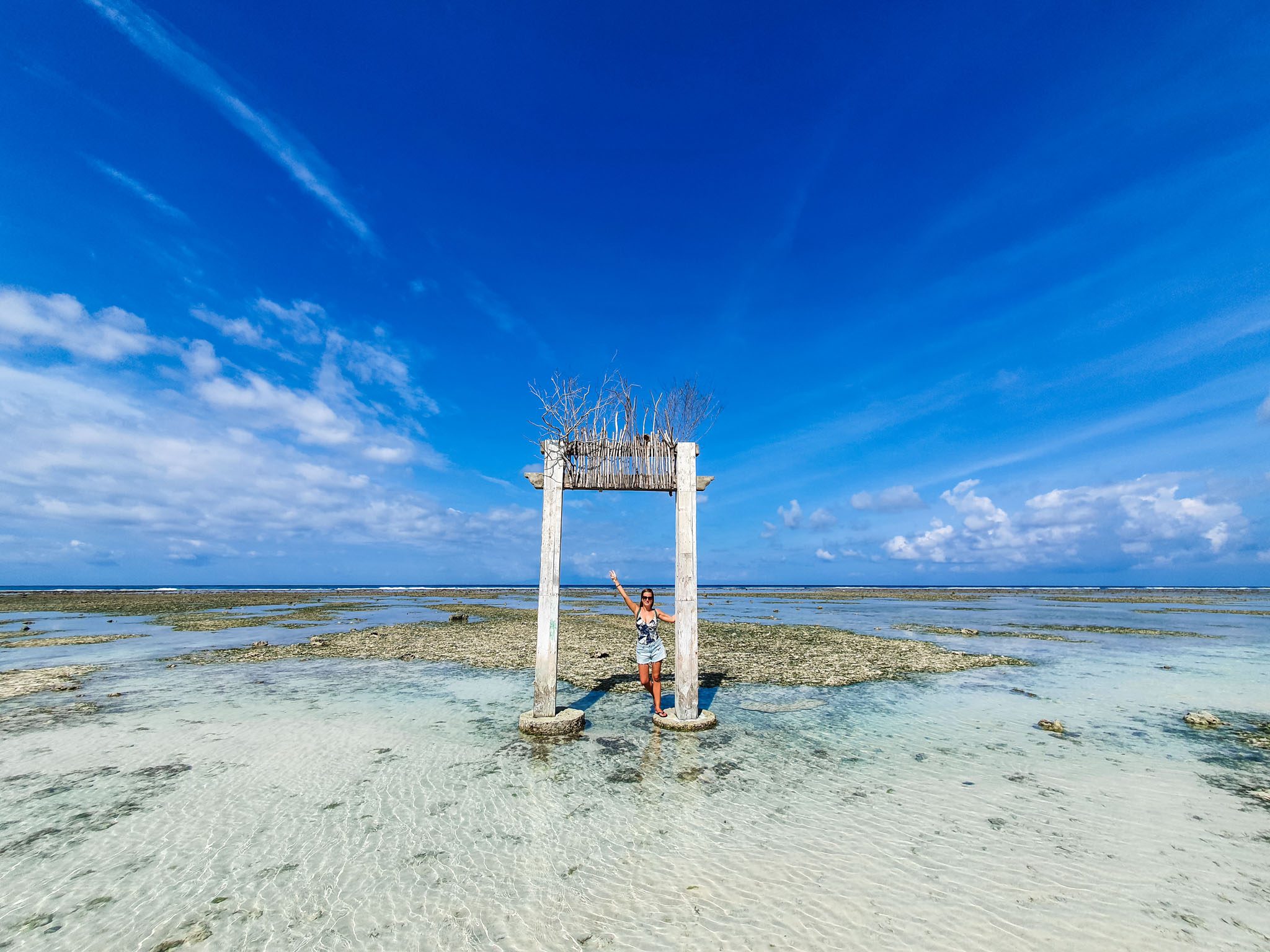 Person posing under a wooden arch on a sunny beach in the Gili Islands.