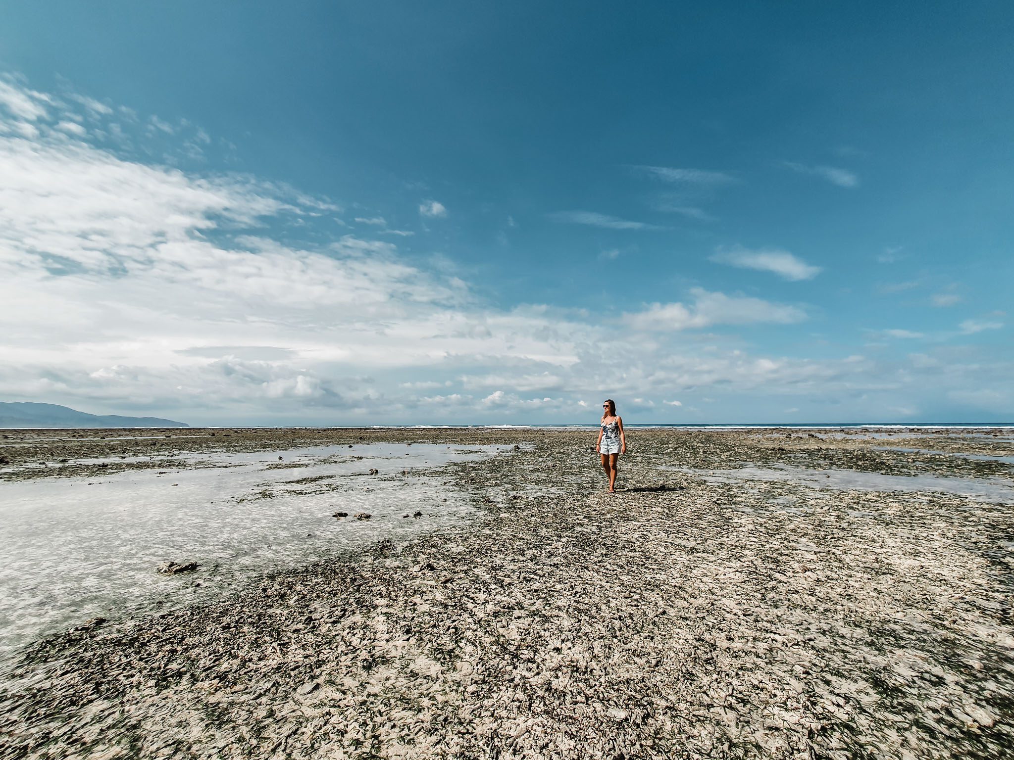 Woman walking along rocky beachfront under blue sky in Gili Islands.