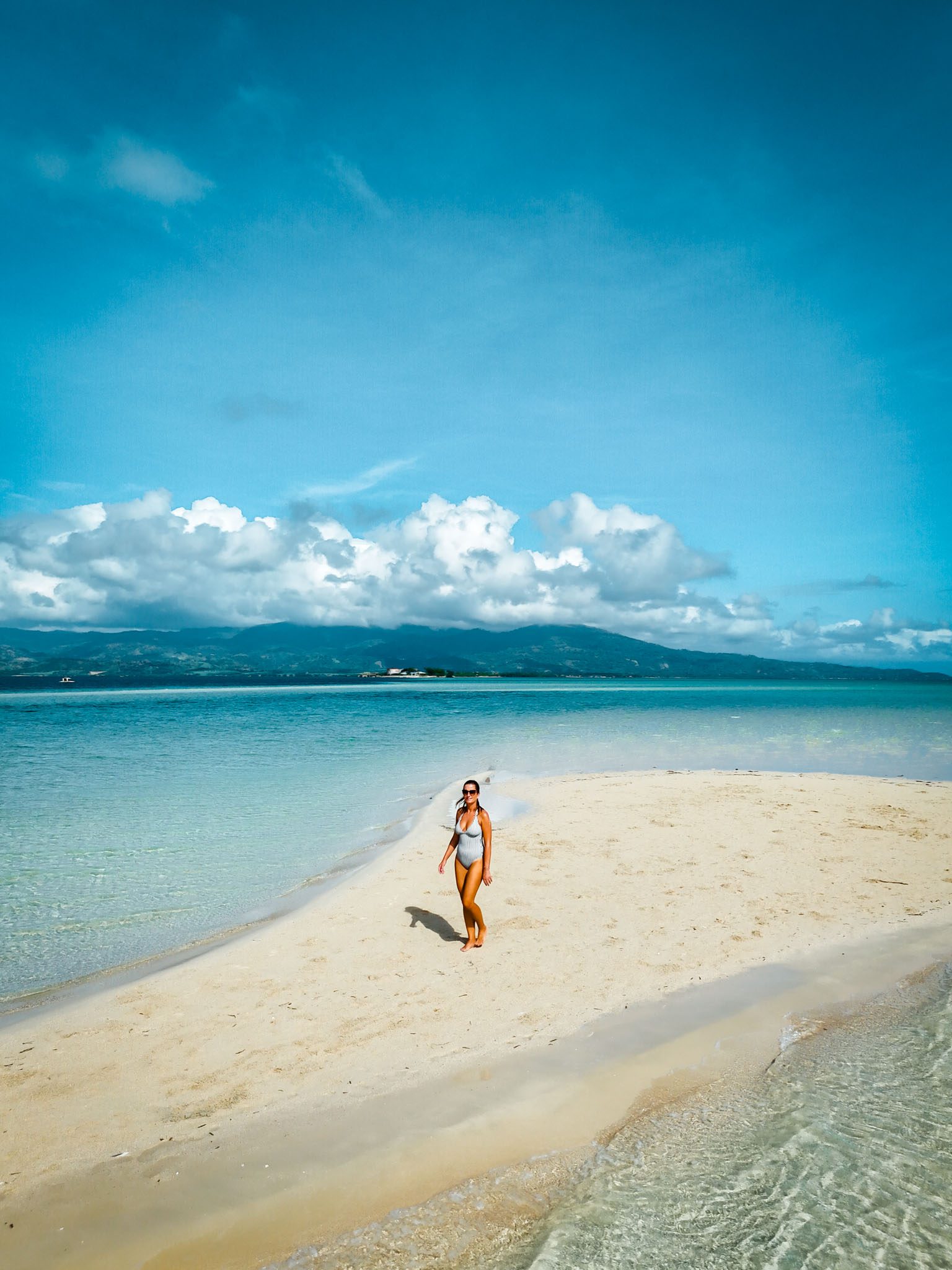 Woman walks on a sandy beach in Dumaguete, Philippines, under a clear blue sky.