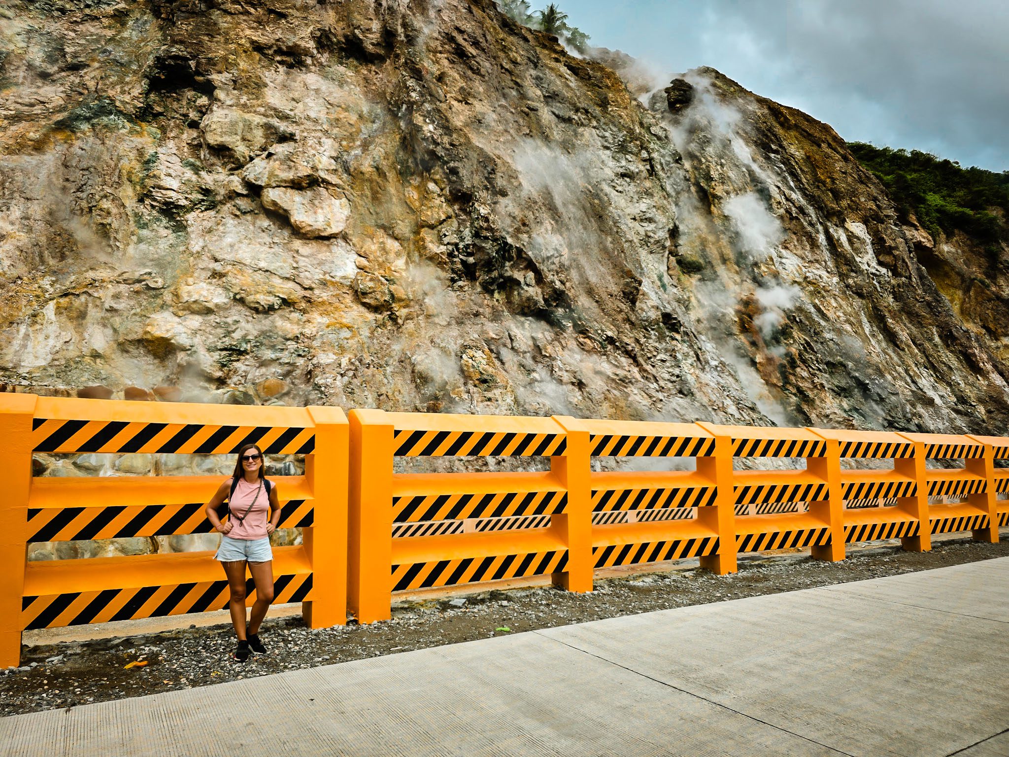 Woman stands by a yellow safety barrier near a steaming cliff in Dumaguete, Philippines.