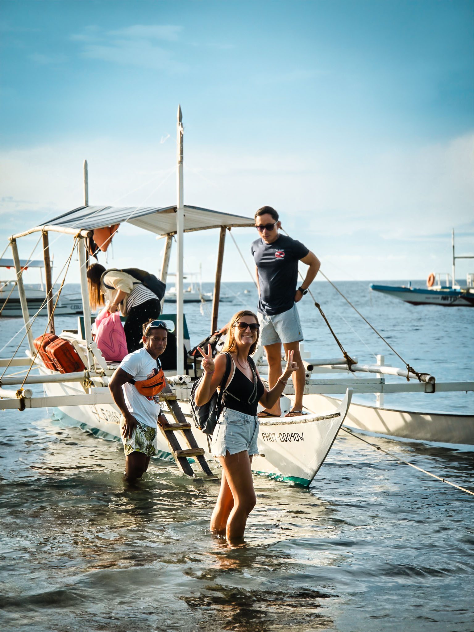 Tourists board a boat in Dumaguete, Philippines, standing in shallow water under a clear sky.