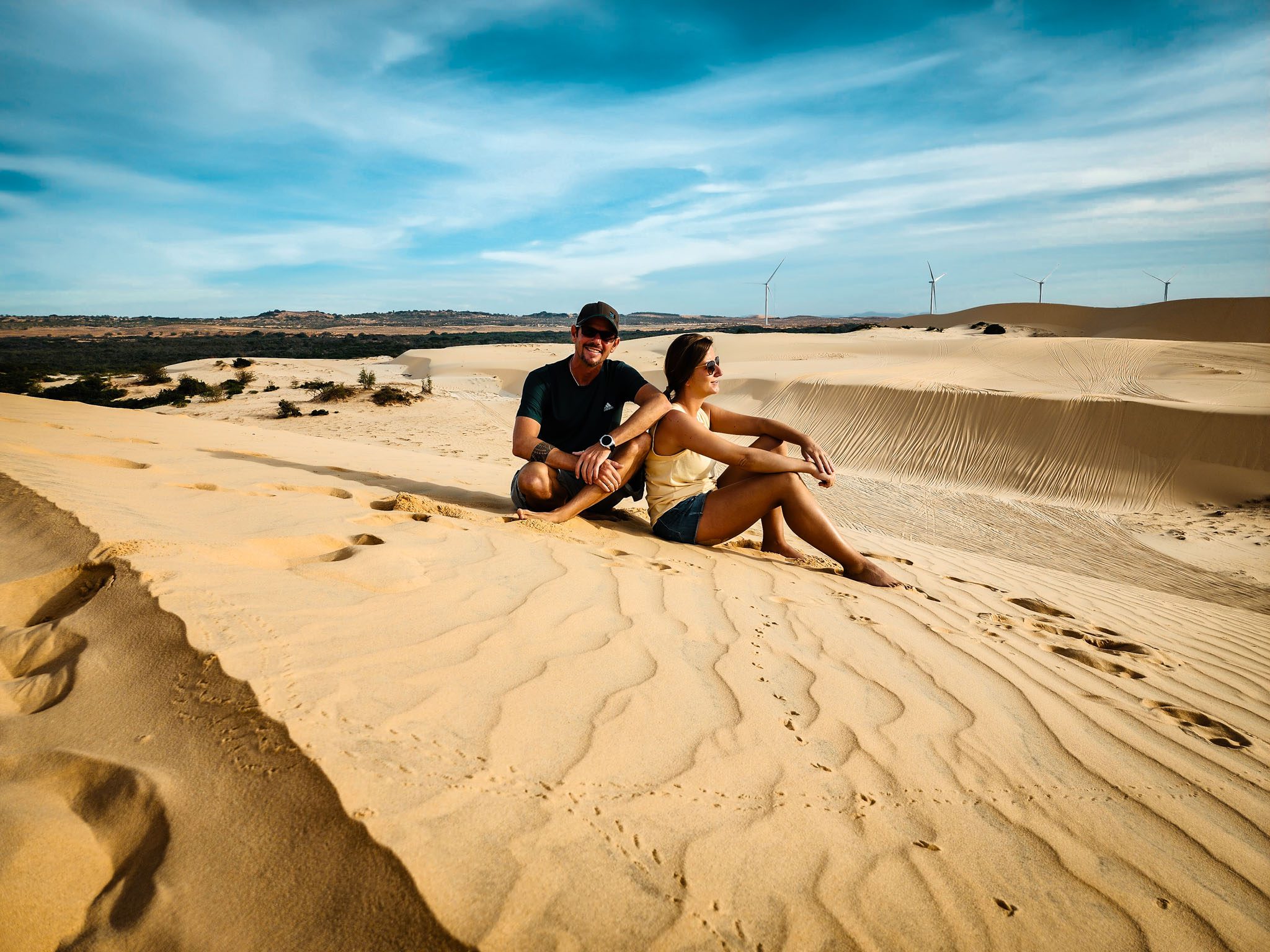 Couple sitting on sand dunes under a blue sky in Mui Ne, Vietnam with wind turbines in view.