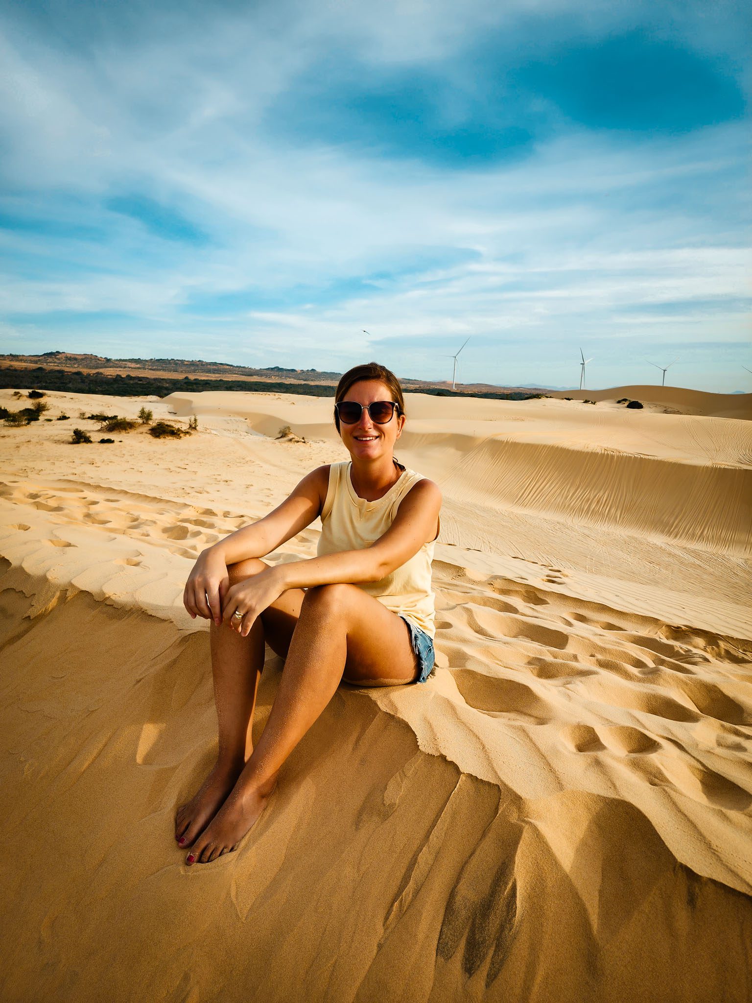 Smiling woman sitting on sand dunes in Mui Ne, Vietnam, under a clear blue sky.