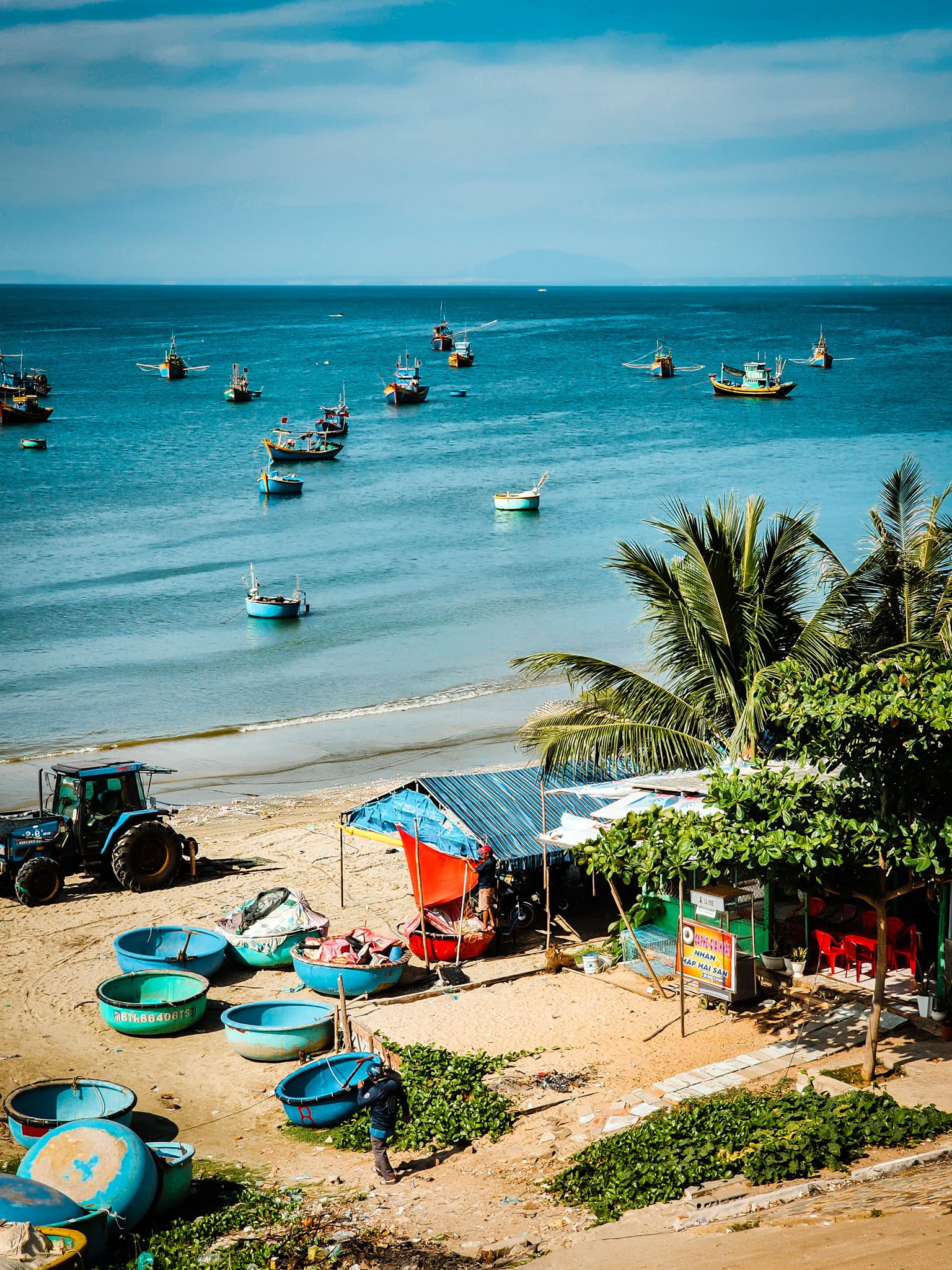 Fishing boats and beachside stalls at Mui Ne, Vietnam under a clear blue sky.