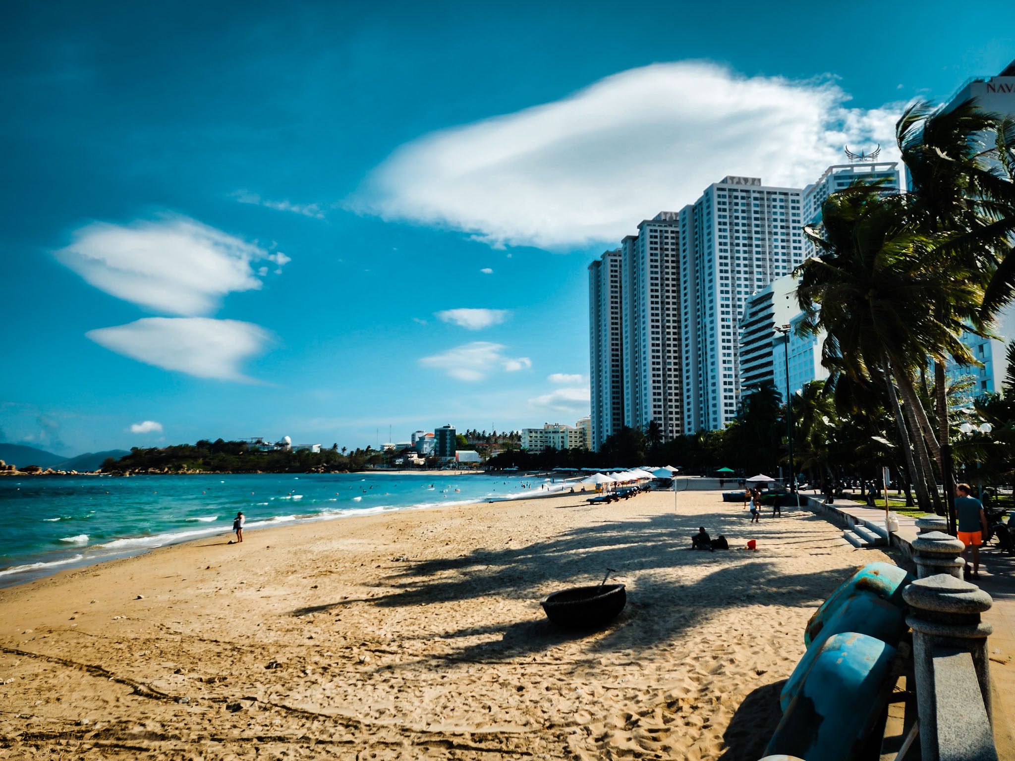 Nha Trang beachfront with high-rise buildings, palm trees, and people enjoying the sandy shore.