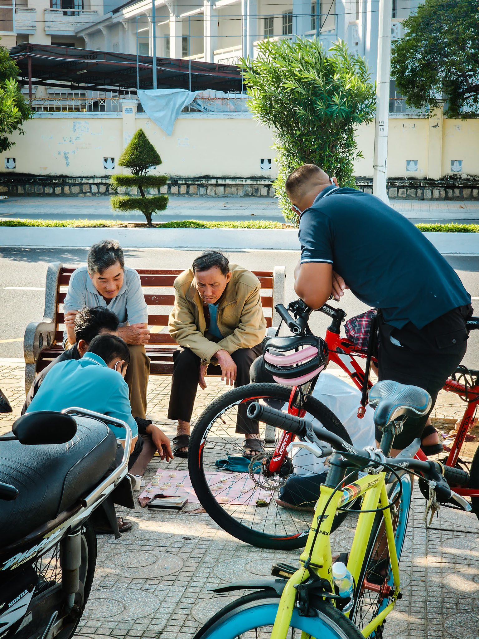 Group playing chess near bikes on a sunny day in Nha Trang, surrounded by greenery.