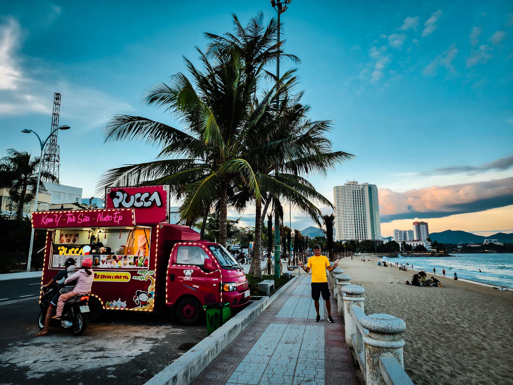 Street food truck and beach promenade at sunset in Nha Trang with palm trees and buildings.