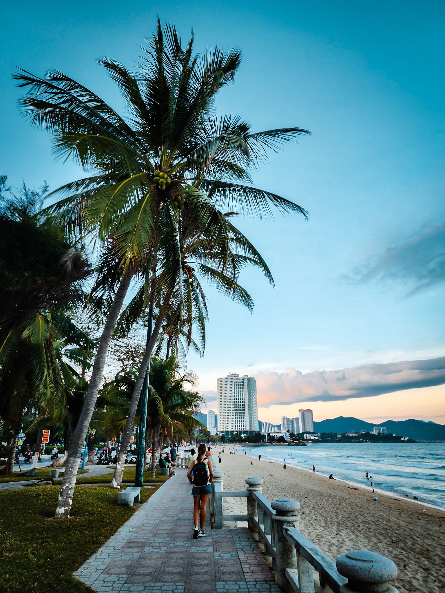 Tourist walking along Nha Trang beach promenade under palm trees at sunset.